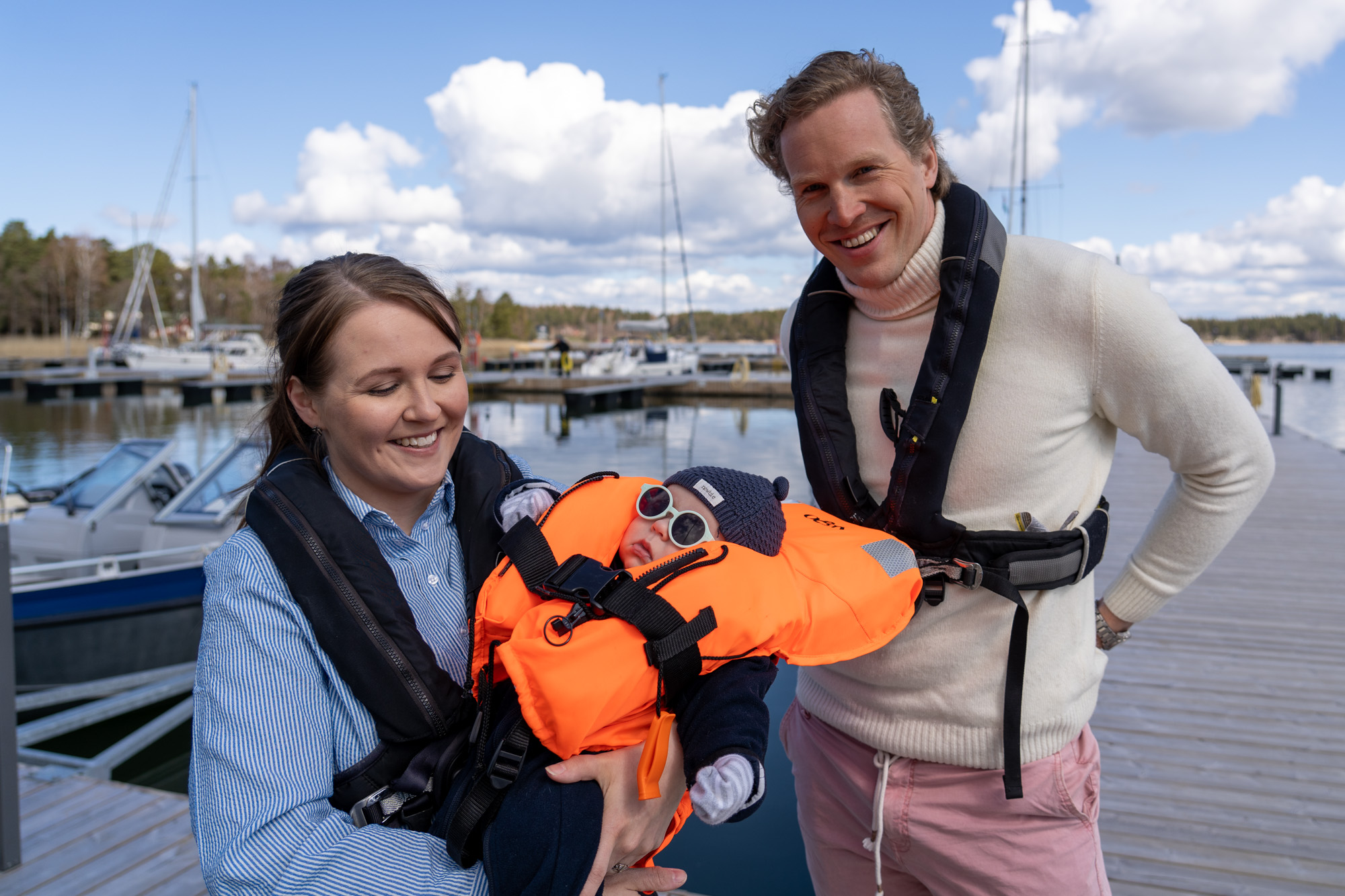 En kvinna och en man står på bryggan med uppblåsbara räddningsvästar på sig. Kvinnan håller en baby i famnen. Babyn har räddningsväst och solglasögon.