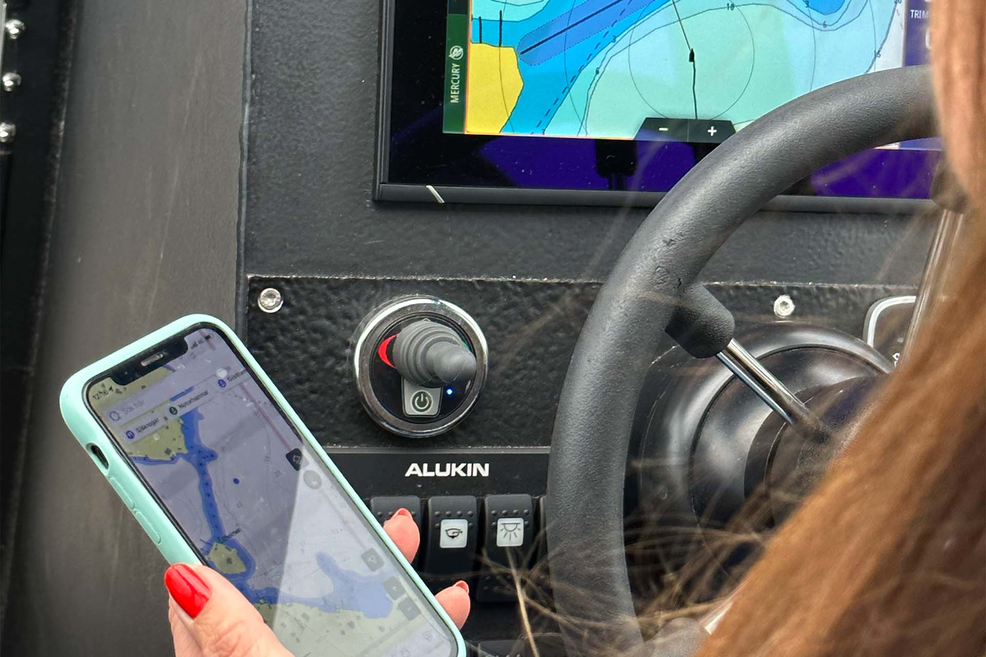 En kvinna sitter vid ratten i en båt med en plotter framför sig och en telefon med navigationsapp i handen.